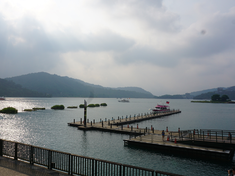Scenic lake with long wooden pier, boats, and distant mountains under a cloudy sky.